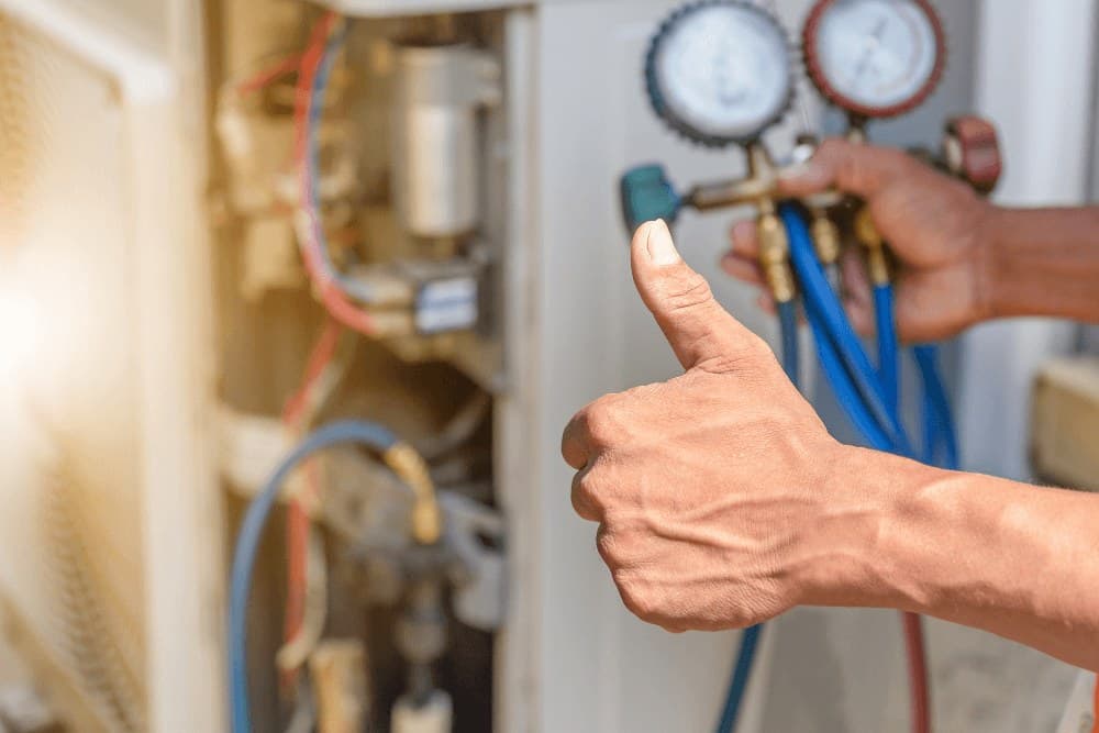 Technician giving a thumbs-up while holding refrigerant hoses and a manifold gauge set at an outdoor air conditioner unit