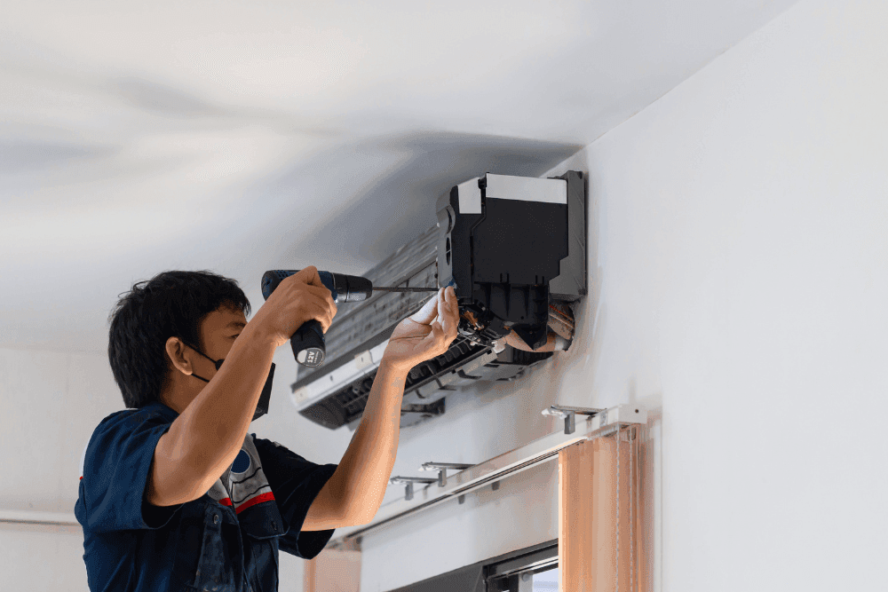 Technician in a face mask using a cordless drill on a split air conditioner indoor unit removed from the wall