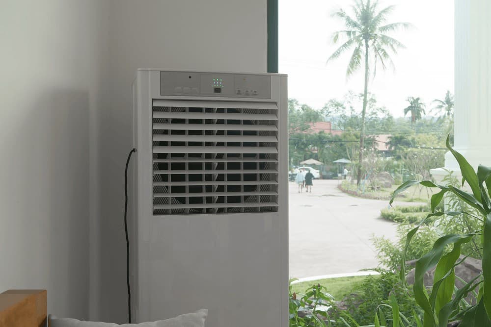 White portable evaporative air cooler standing on a tiled floor beside a window with a view of palm trees, positioned for use in a home living space