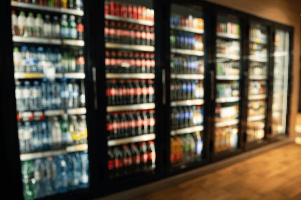 Row of illuminated commercial glass-door beverage coolers stocked with drinks in a retail or restaurant setting, the kind of commercial refrigeration unit that needs regular servicing