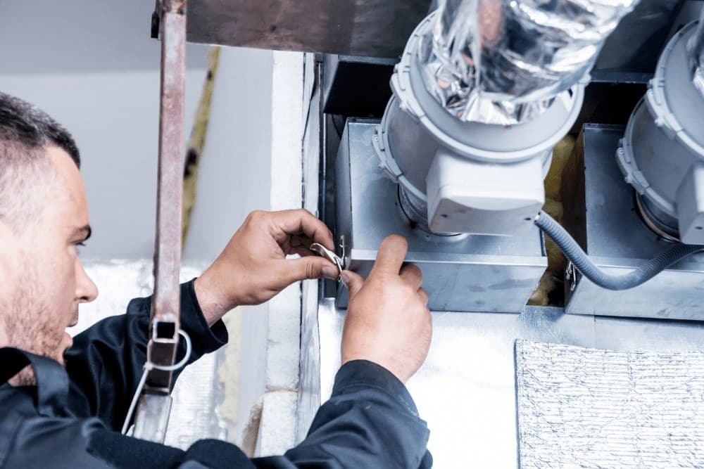 Technician's hands securing the metal housing of an in-line fan and flexible aluminium ducting during an air duct service