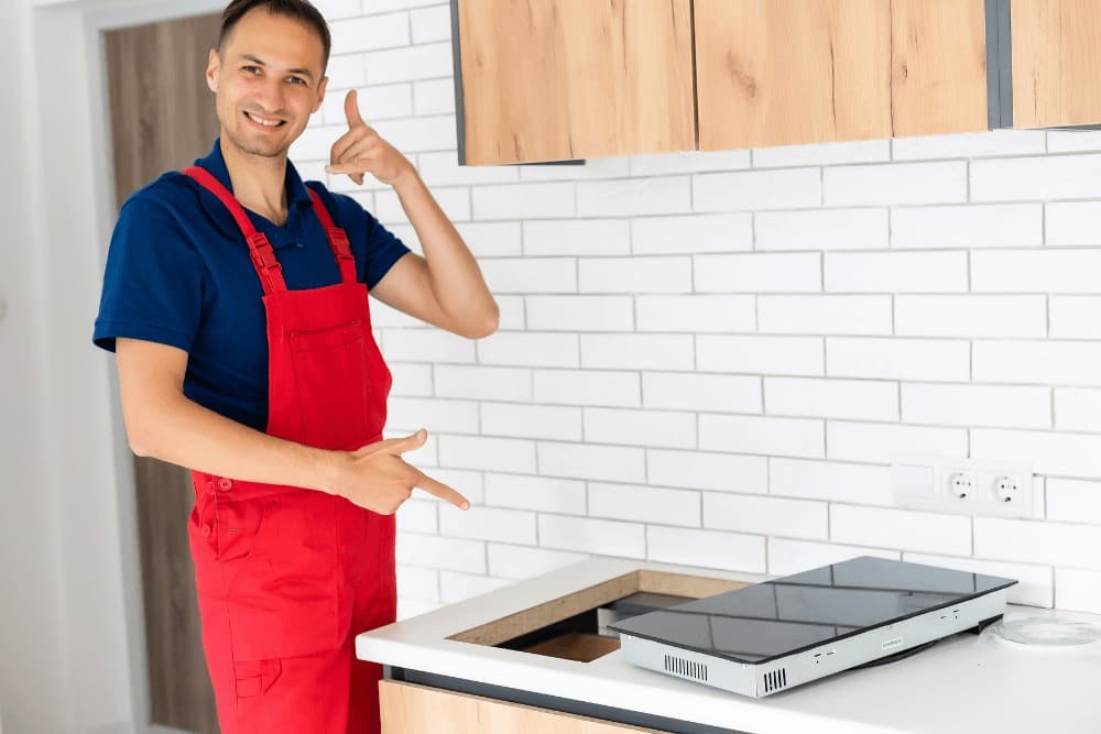 Smiling technician in red overalls pointing at a newly installed induction hob set into a kitchen worktop, beside a white-tiled wall and wooden cabinets