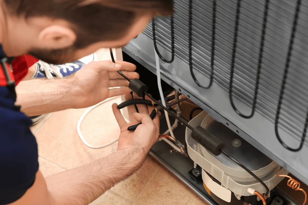 One Click Technicians technician's hands working on a refrigerator's rear compressor compartment, connecting wiring beside the condenser coil during a repair in Abu Dhabi