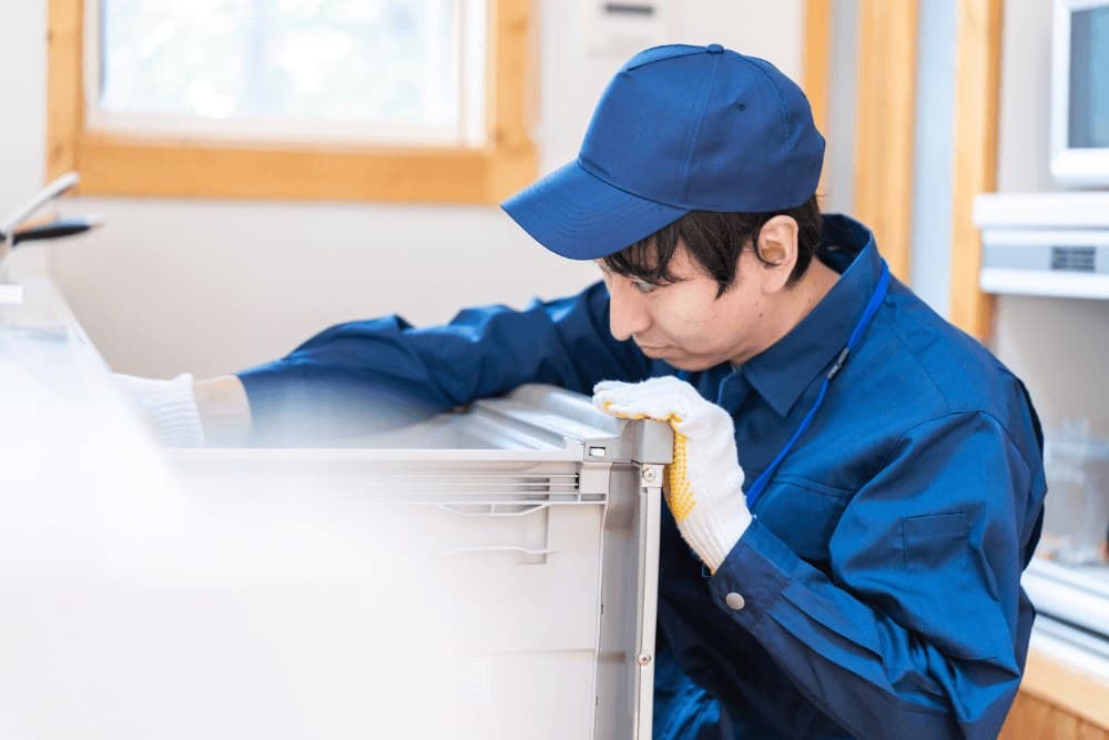 Technician in a blue cap and uniform leaning in to check the door edge and seal of a white refrigerator in a bright kitchen