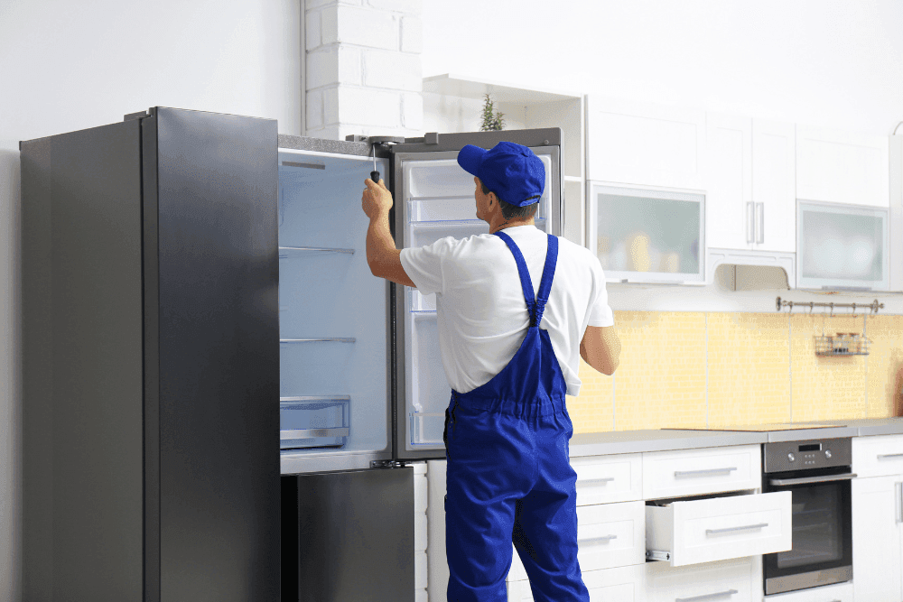 Technician in blue overalls checking the inside of an open French-door refrigerator in a modern kitchen