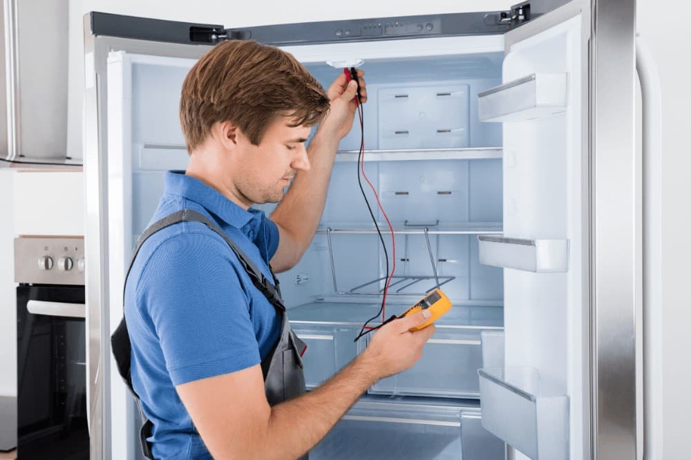Technician in a blue polo shirt and tool suspenders standing in front of an open refrigerator, testing the interior with a multimeter