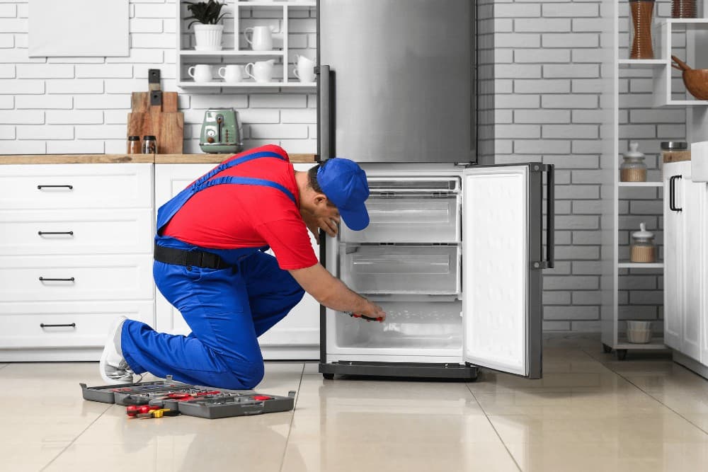 Technician in red overalls and a blue cap kneeling in front of an open upright refrigerator-freezer, working on its internal panel with a toolbox open on the floor