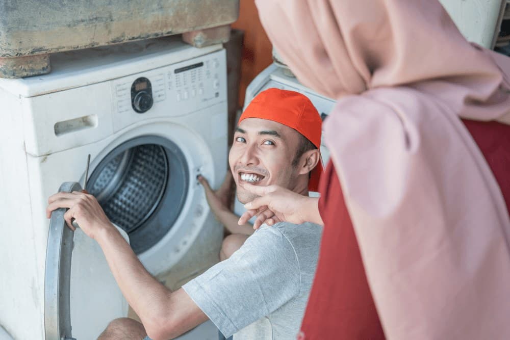 Smiling technician in a red cap checking the door seal of a front-loading washing machine as a homeowner in a headscarf points to it
