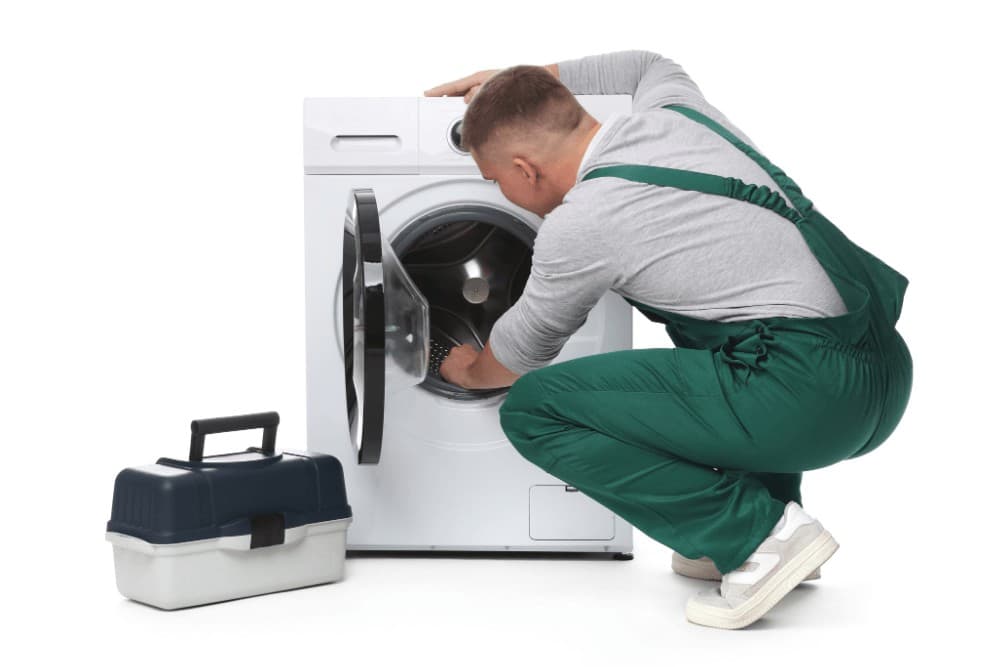Technician in green overalls kneeling in front of an open front-loading washing machine, working on its drum during a repair