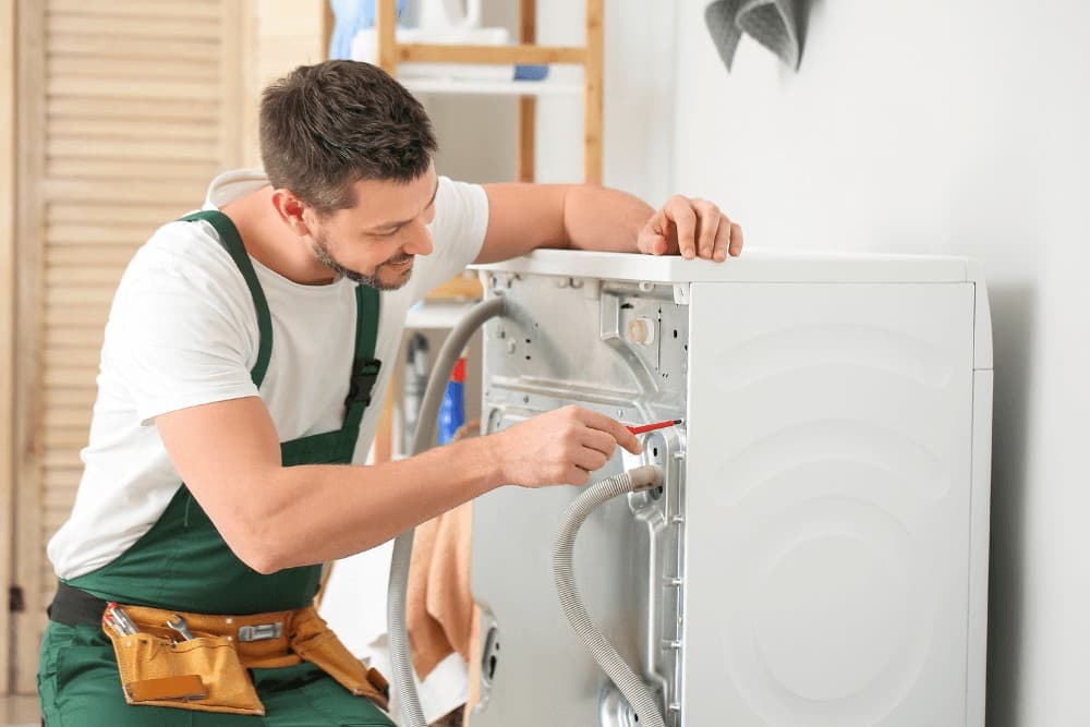 Technician in green overalls working on the rear panel of a washing machine, checking the hose connections during a repair