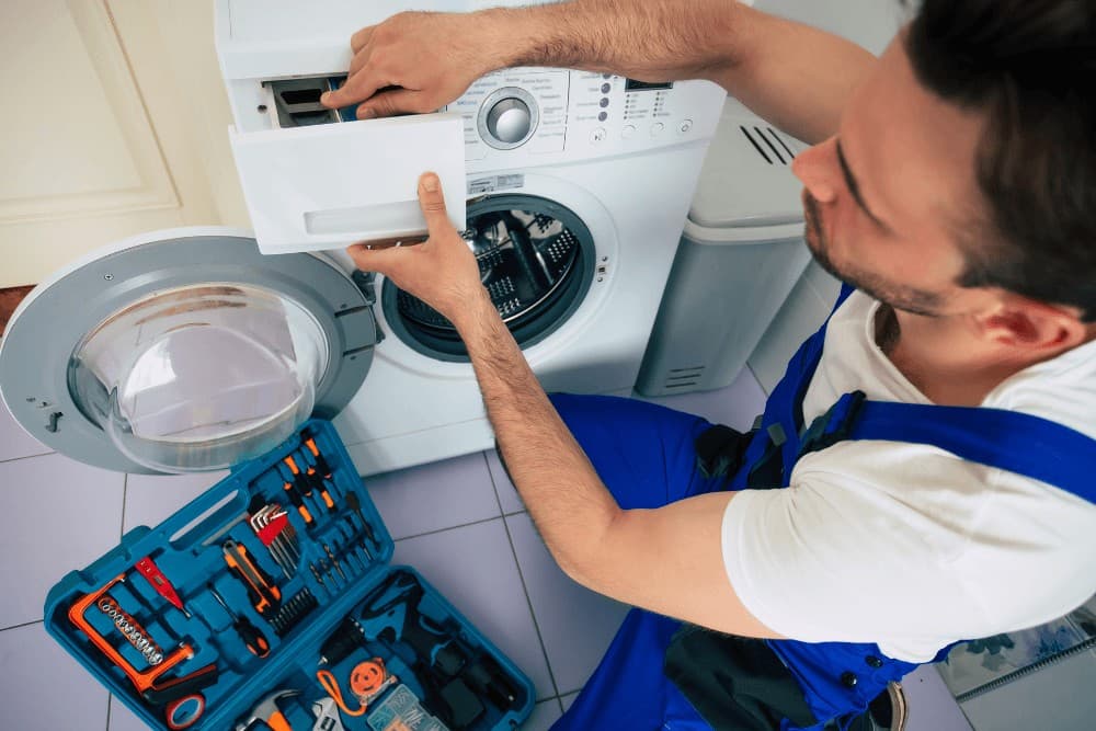 Technician pulling out the detergent drawer of a front-loading washing machine, with a toolbox open on the floor beside him