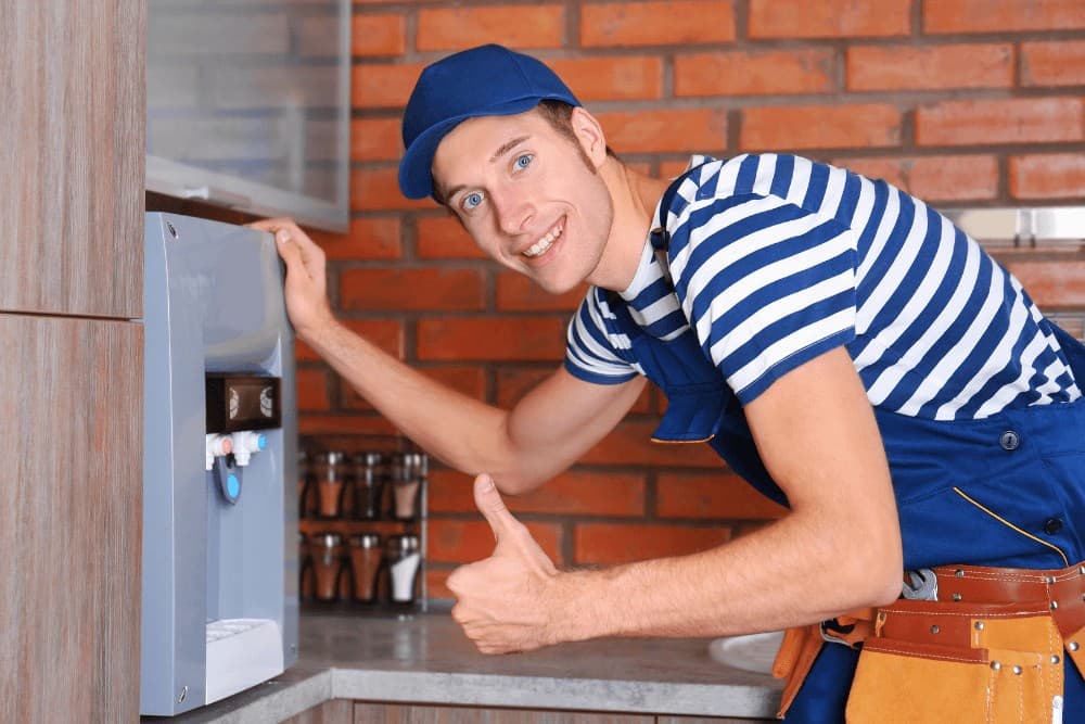Technician in a striped shirt and cap giving a thumbs-up beside a wall-mounted water dispenser with its cold and hot taps visible, confirming the repair is complete in a kitchen