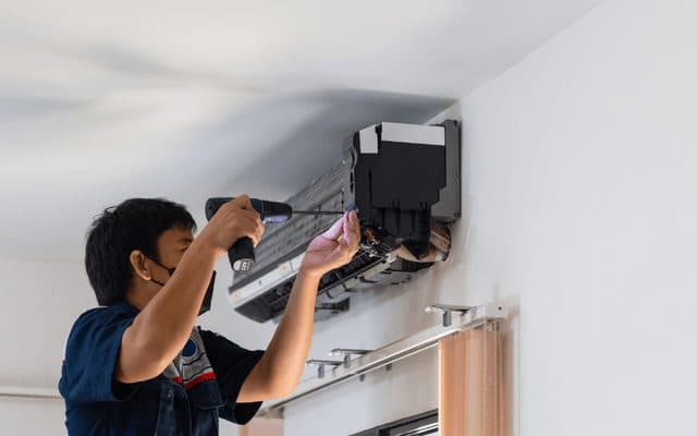 Technician in a face mask using a cordless drill on a split air conditioner indoor unit removed from the wall
