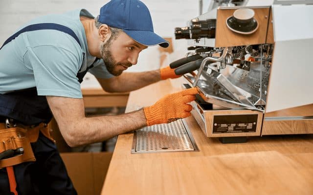 Technician in orange gloves servicing a commercial espresso machine on a café counter