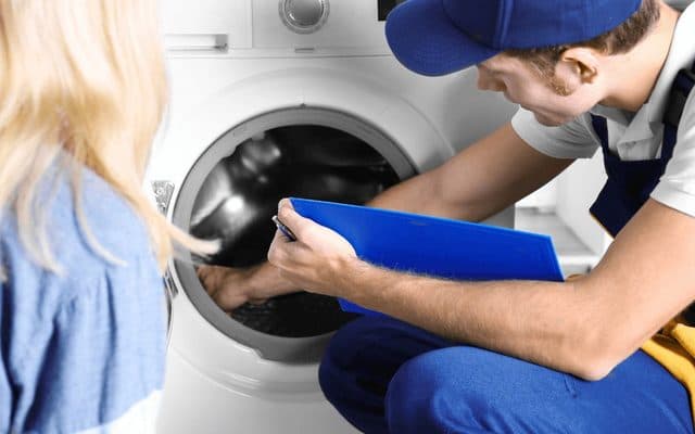 Technician in a blue cap writing on a clipboard while inspecting the drum of a washing machine as the homeowner looks on