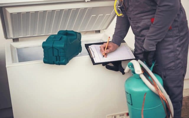 Technician in gray overalls with safety goggles at his collar writing on a clipboard beside an open chest freezer, with a teal toolbox on the lid and a refrigerant cylinder and hoses at the front
