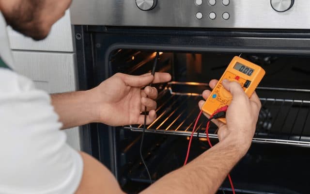 Hands holding a multimeter probe against the heating element inside an open built-in oven