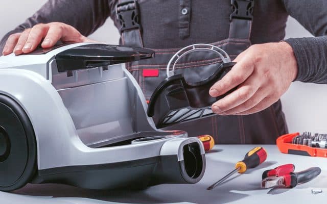 Hands reassembling a gray vacuum cleaner's dust canister and motor housing on a workbench, with a toolbox and spare parts laid out beside the unit