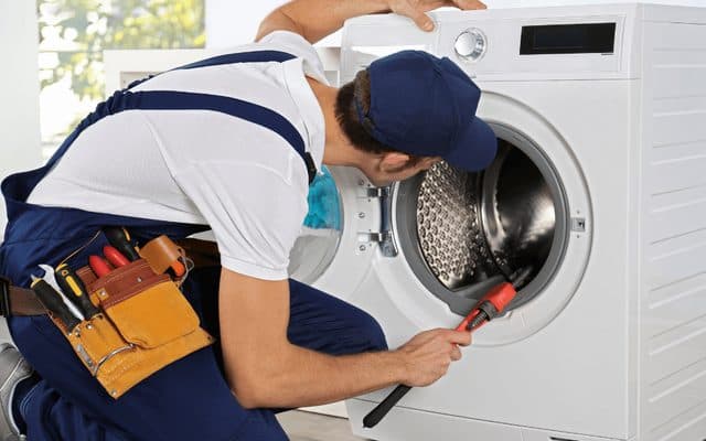 Technician in blue overalls kneeling beside a front-loading washing machine, using a tool inside the open drum