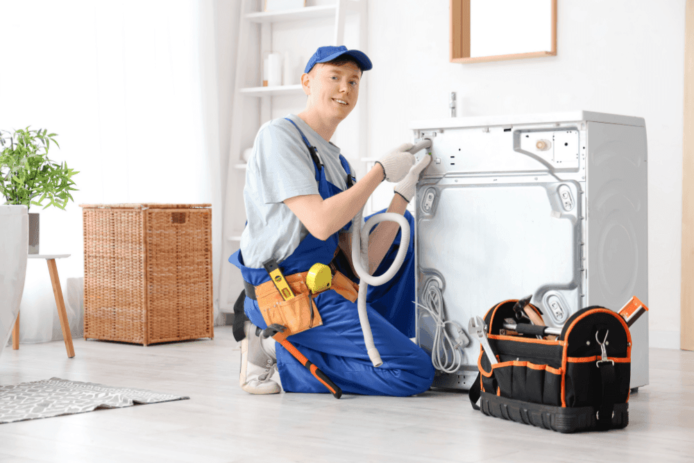 Smiling technician kneeling behind a washing machine, connecting a hose, with a tool bag on the floor