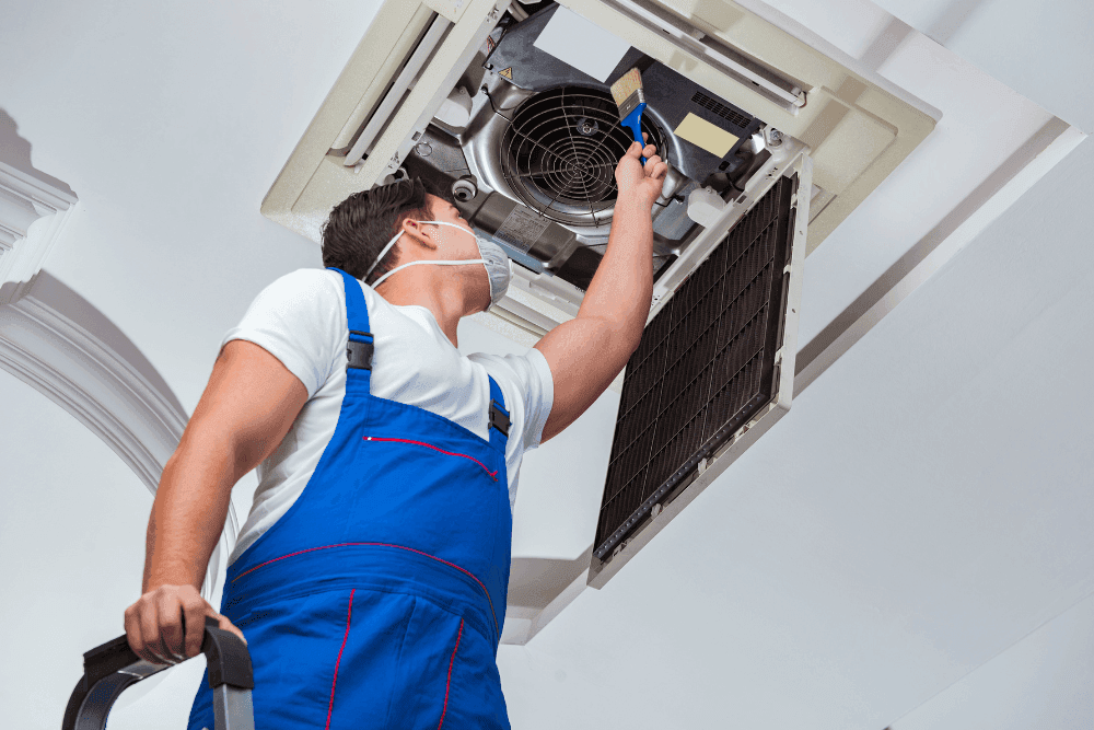 Technician in a face mask on a ladder cleaning the fan and filter of a ceiling cassette air conditioner