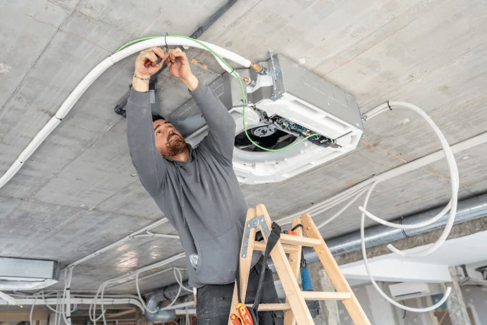 Technician on a stepladder fitting a ceiling cassette air conditioner and running its cables across a concrete ceiling