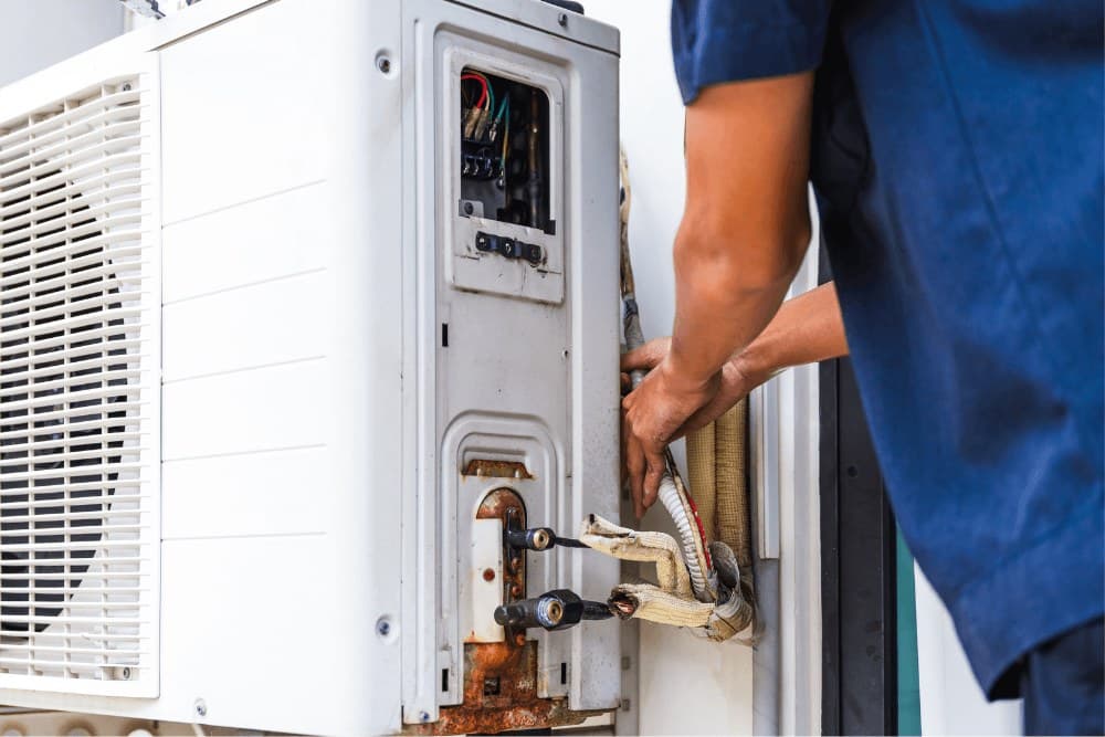Technician in a blue shirt working on the wiring and refrigerant fittings of an outdoor air conditioner unit