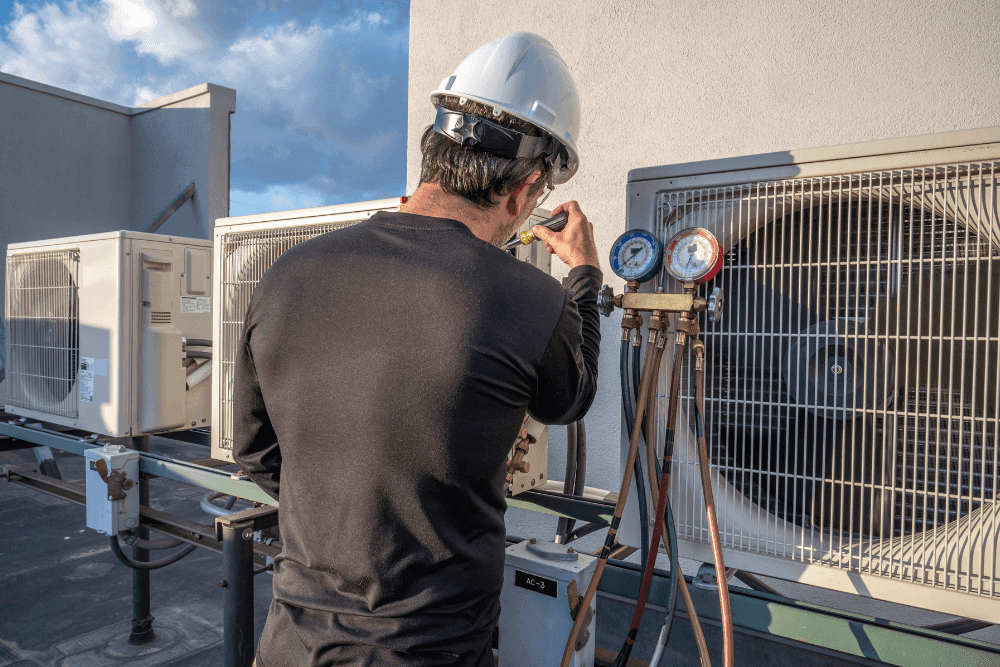 Technician in a hard hat connecting pressure gauges to rooftop air conditioning condenser units