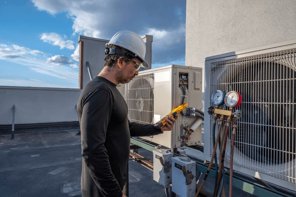 Technician in a hard hat and safety glasses holding a clamp meter beside a manifold gauge set connected to a rooftop condenser unit during a VRV diagnosis