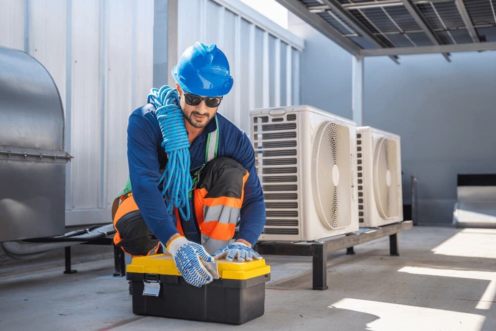 Technician in a blue hard hat and gloves kneeling over a toolbox beside rooftop air conditioner condenser units