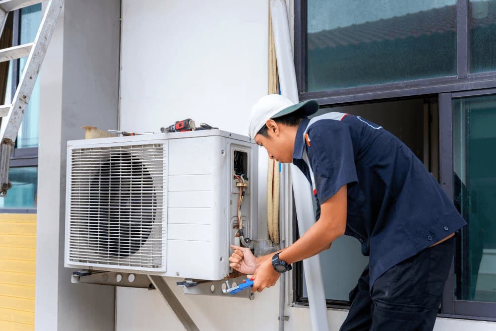 Technician in a hard hat and navy shirt connecting the pipework of a wall-bracket-mounted outdoor air conditioner unit