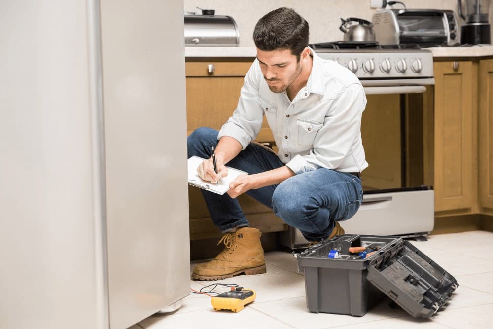 Technician crouching in a kitchen and writing on a clipboard beside a refrigerator, with an open toolbox and a multimeter on the floor