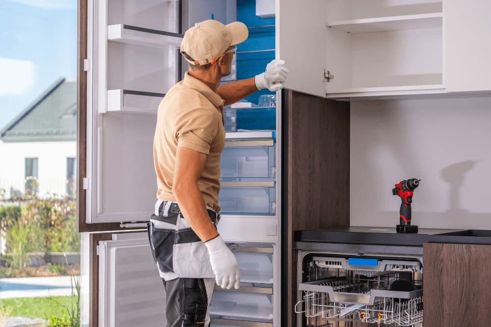Built-in kitchen with an integrated dishwasher, its lower rack pulled out, next to a technician working on the neighbouring fridge column, the kind of integrated Bosch and Siemens installation we service