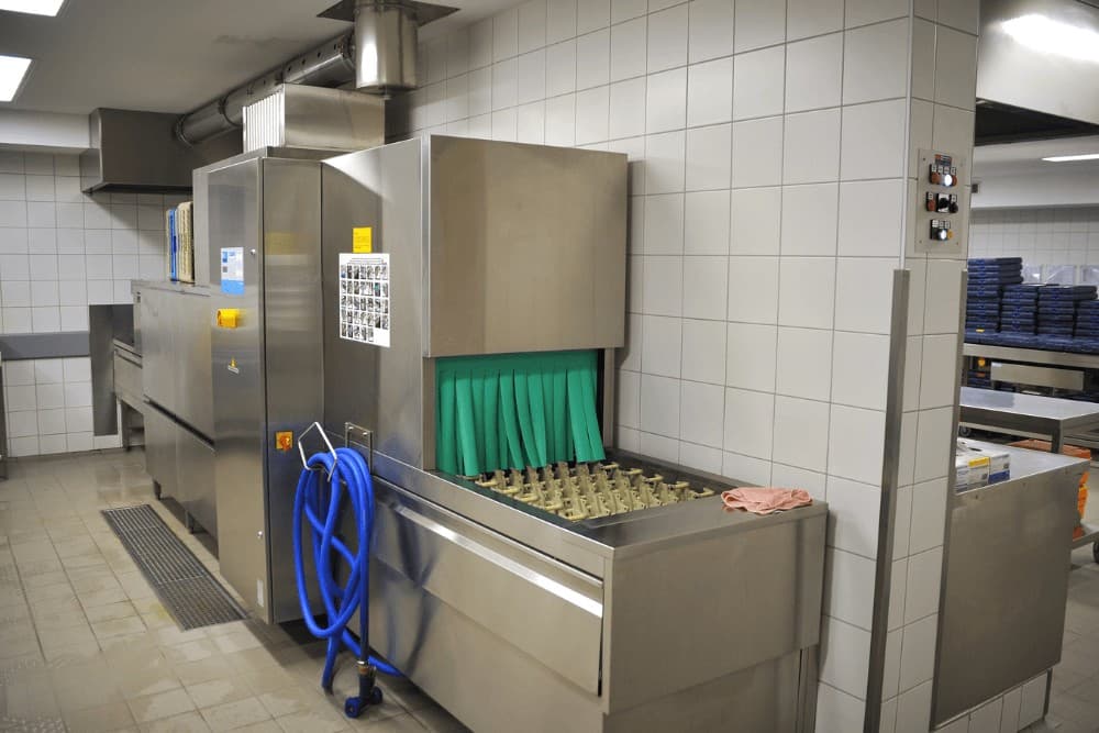 Commercial stainless steel dishwasher tunnel unit in a restaurant back-of-house kitchen, with its plastic curtain strips and connected hose visible at the entry point