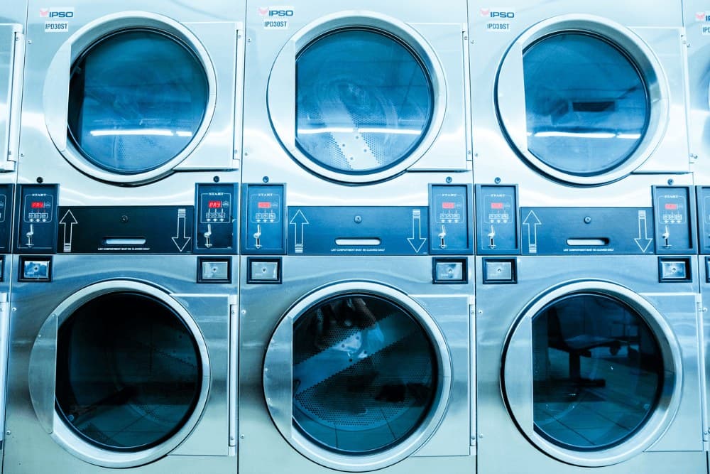 Stacked coin-operated dryers above washing machines in a laundromat, with control panels between the rows