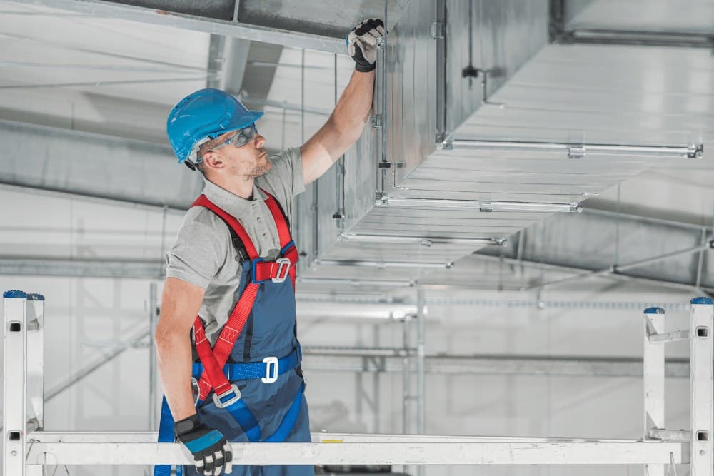 Technician in a hard hat and safety harness on a scaffold, working on a large overhead metal air duct