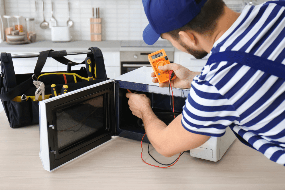 Technician testing the wiring of an open microwave with a multimeter on a kitchen counter, toolbox beside it