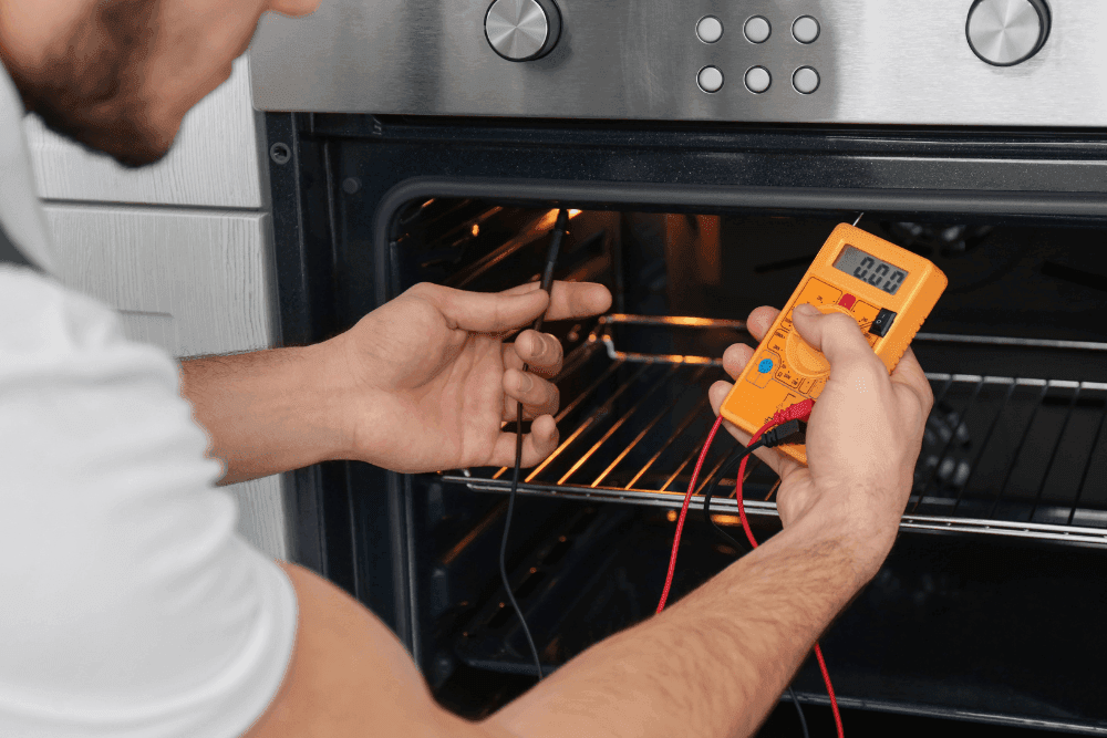 Hands holding a multimeter probe against the heating element inside an open built-in oven