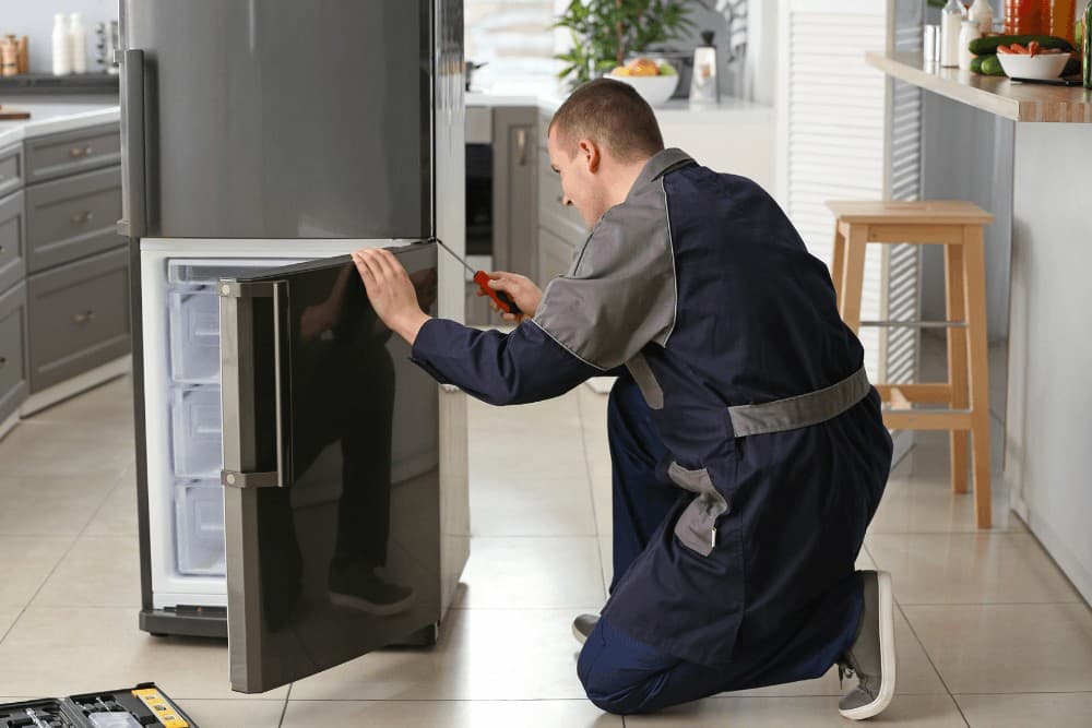 Technician in a black work apron kneeling in front of an open refrigerator, adjusting the door hinge alignment during a repair in a kitchen