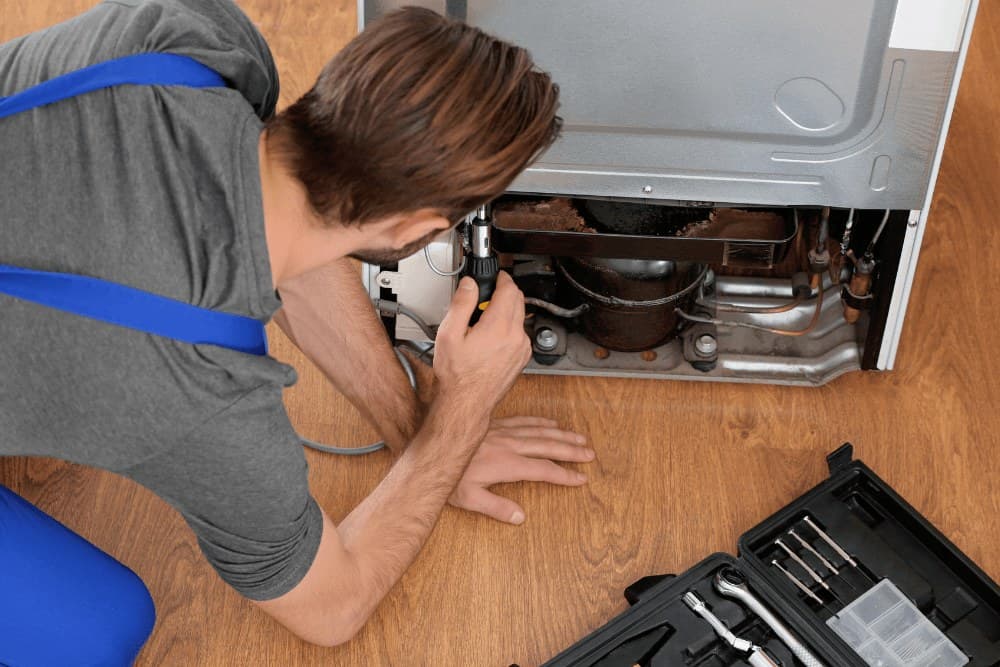 Technician in blue overalls kneeling on the floor and shining a torch at the compressor compartment at the base of a refrigerator, the same sealed-system check done on wine coolers
