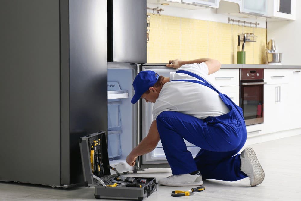 Technician in blue overalls kneeling in front of an open refrigerator in a kitchen, with an open toolbox on the floor