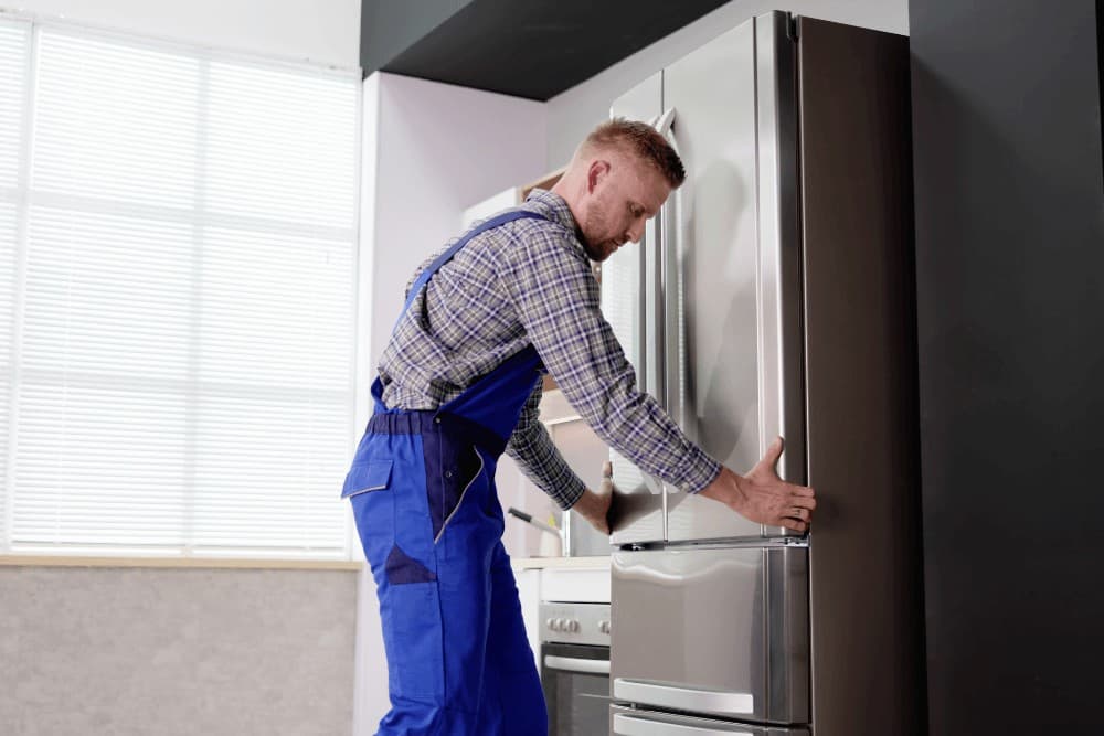 Technician in a plaid shirt and dark overalls working on the side panel of a tall refrigerator beside a window, during a repair