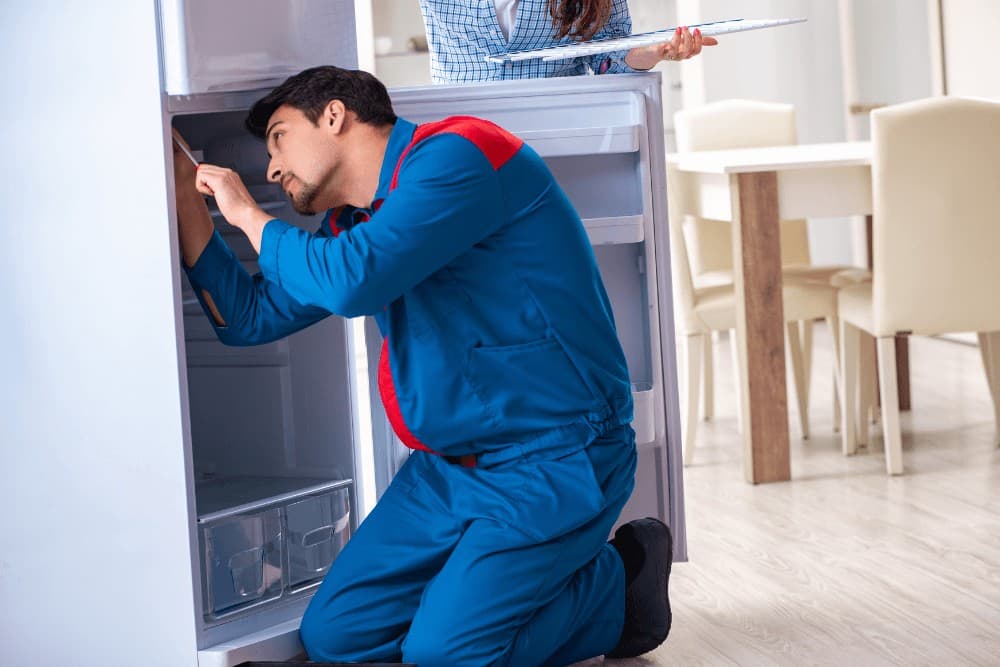 Technician in red and blue overalls kneeling in front of a white refrigerator, working on its open front panel with a dining area visible behind