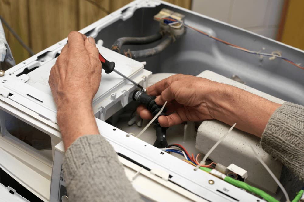 Hands working inside the open rear panel of a washing machine, checking the internal wiring during a repair