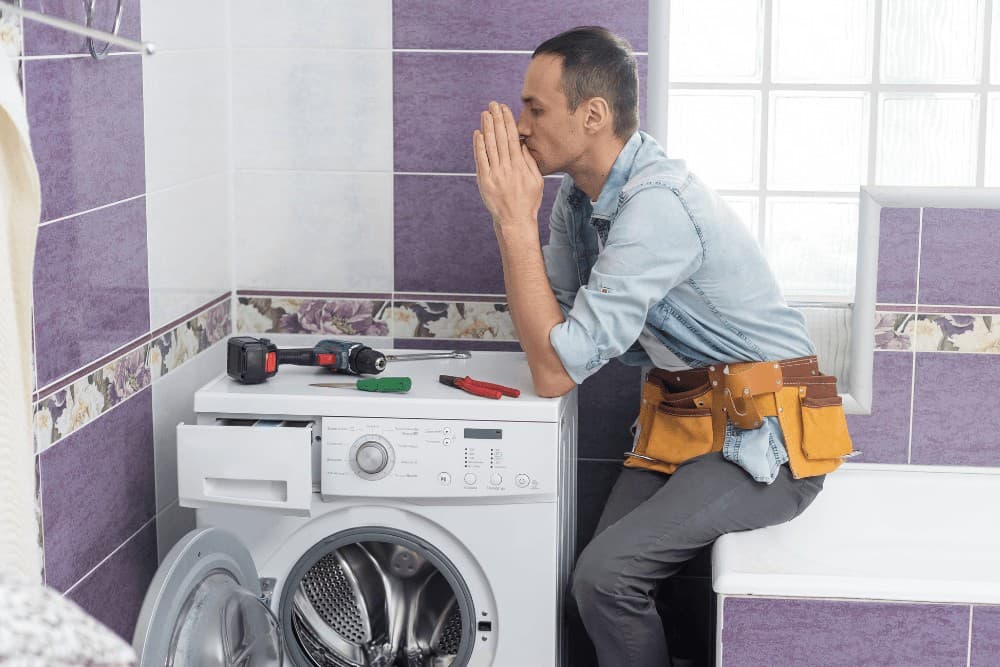 Technician with a tool belt resting his chin on his hands beside a washing machine in a purple-tiled bathroom, tools on top of the machine