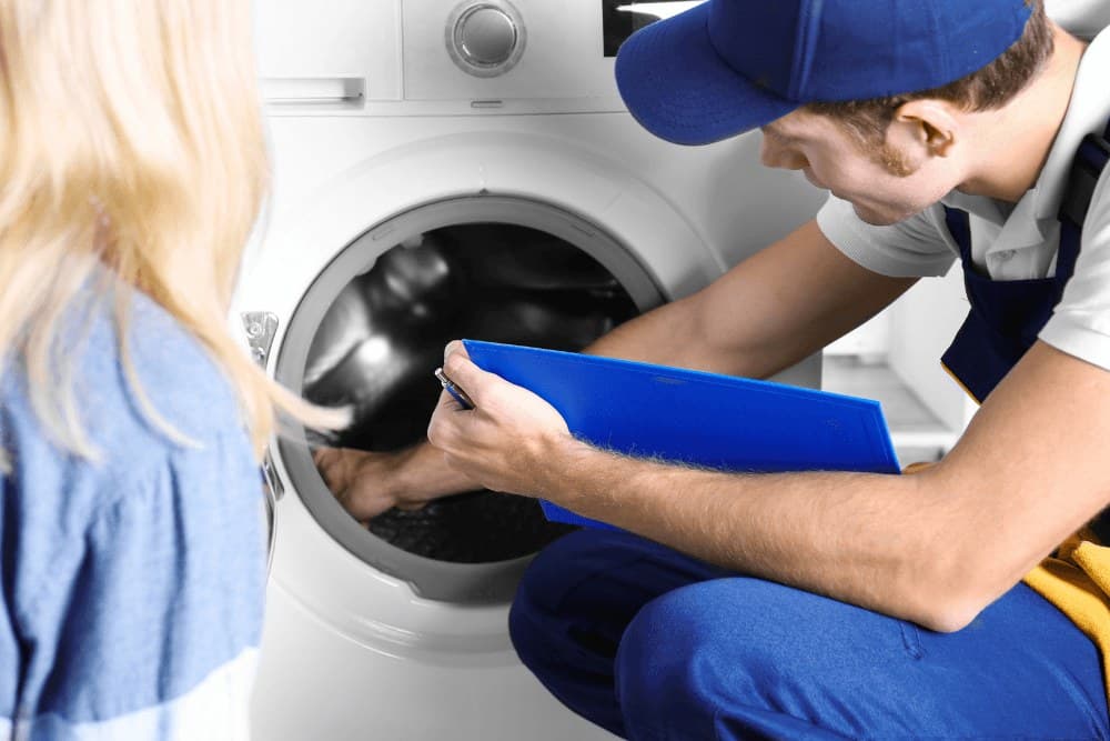 Technician in a blue cap writing on a clipboard while inspecting the drum of a washing machine as the homeowner looks on