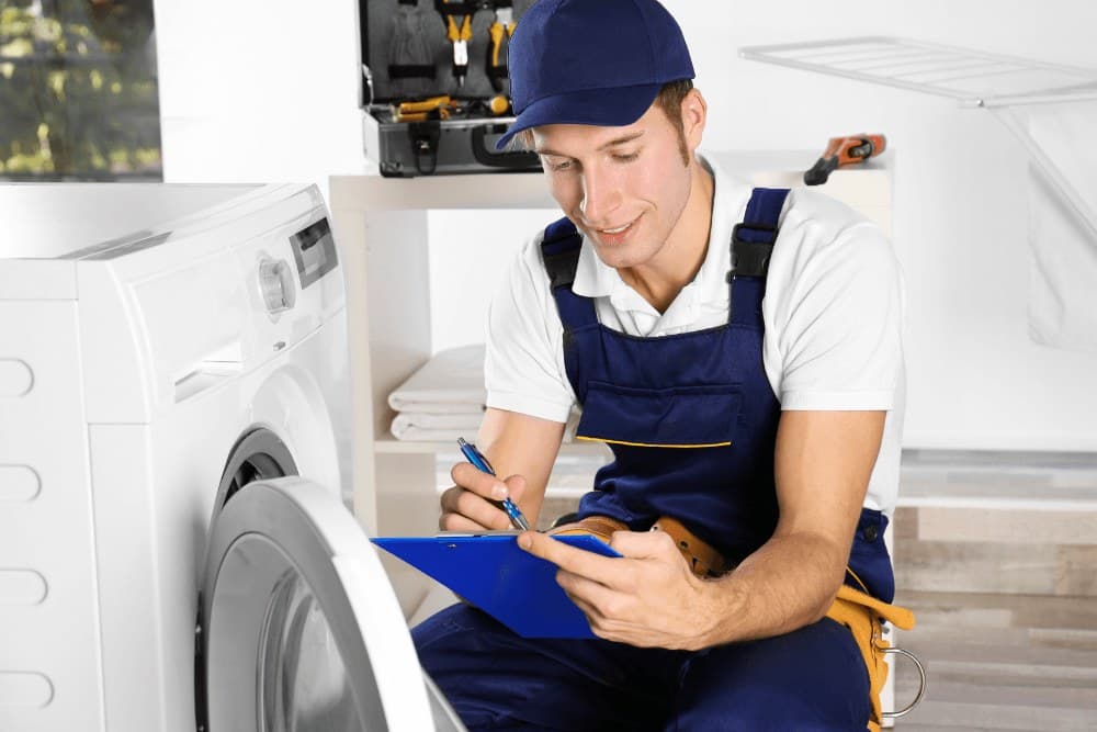 Smiling technician in blue overalls sitting beside a washing machine and writing on a clipboard during a Toshiba washing machine repair visit in Abu Dhabi