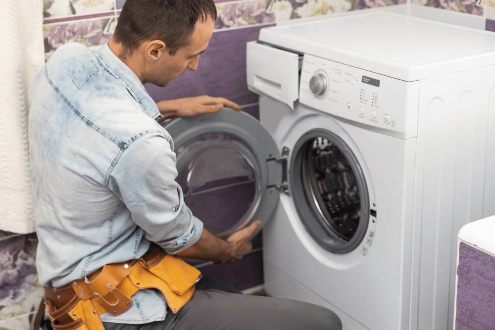 Technician in a denim shirt and tool belt opening the door of a front-loading washing machine