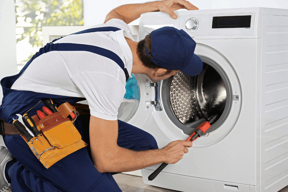 Technician in blue overalls kneeling beside a front-loading washing machine, using a tool inside the open drum