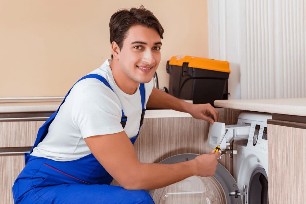 Smiling technician in blue overalls crouching beside a washing machine, holding a spanner at the water connection